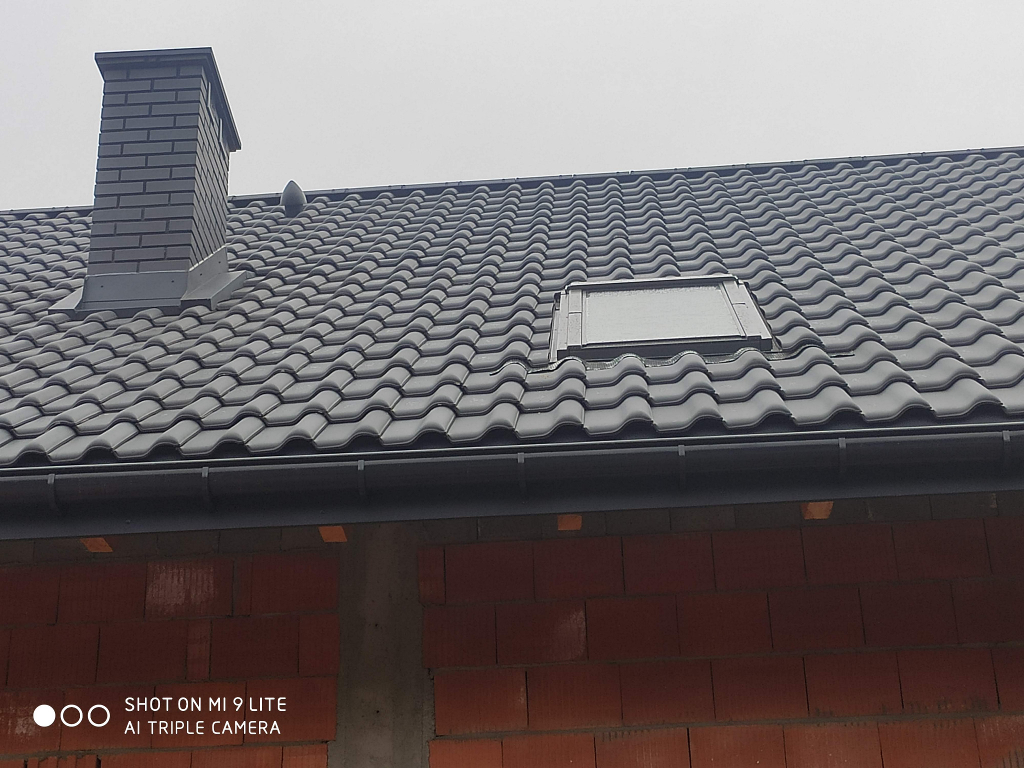 Nowy dach z ciemnej dachówki ceramicznej, okno dachowe i komin z cegły klinkierowej, widok z dołu na tle szarego nieba, ściany budynku w trakcie budowy.