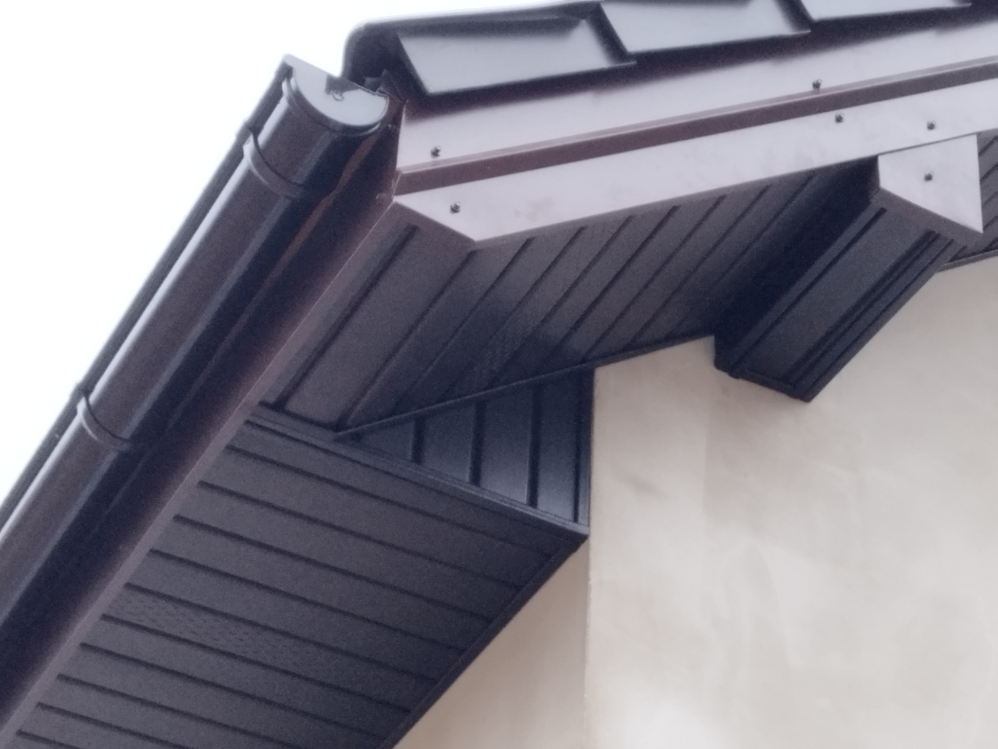 Fragment dachu z brązową blachą trapezową, orynnowaniem i podbitką, widok z dołu na tle jasnej ściany.