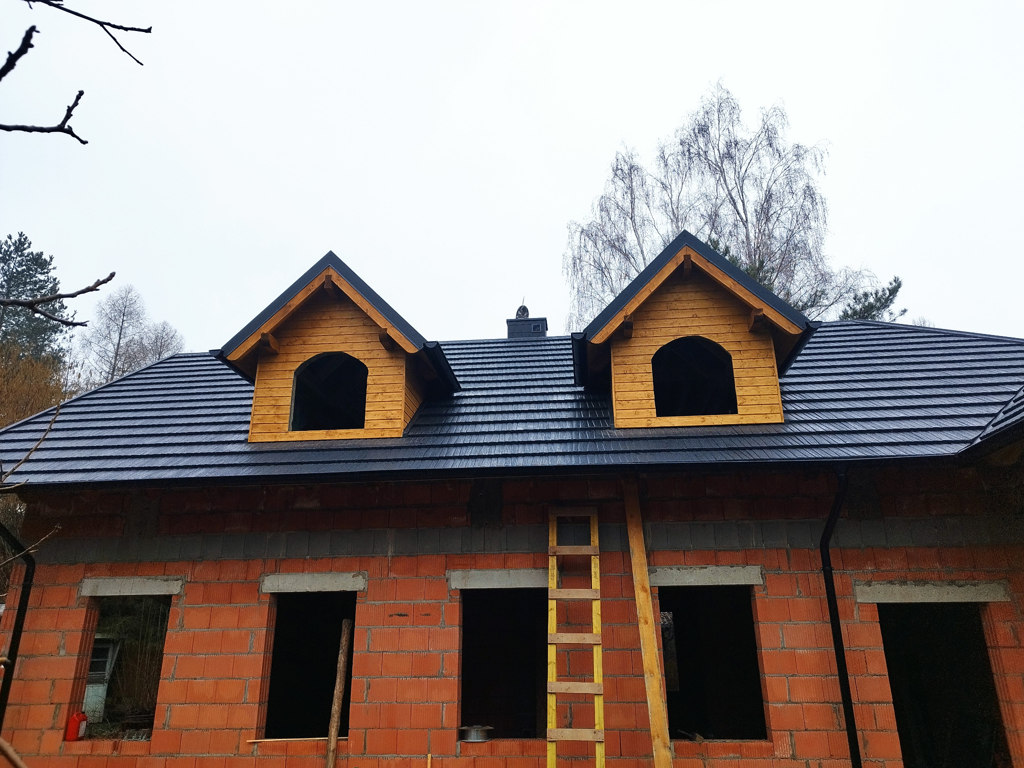 Surowy stan domu z cegły z zamontowanym, ciemnym dachem z dwoma drewnianymi lukarnami i drabiną opartą o ścianę.