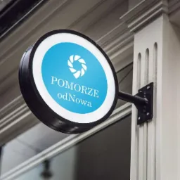 Elegancki, okrągły szyld reklamowy z podświetleniem, w kolorach błękitu i bieli, przymocowany do fasady budynku z oknami, prezentujący logo i nazwę 'POMORZE odNowa'.