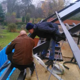 Dwóch mężczyzn w trakcie montażu konstrukcji dachu z metalowych profili, widoczne narzędzia i materiały izolacyjne, w tle zieleń drzew i zabudowania.