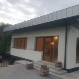 Nowoczesny dom z jasną elewacją i ciemnoszarą, płaską blachą na dachu, widoczne schody wejściowe i fragment podjazdu.