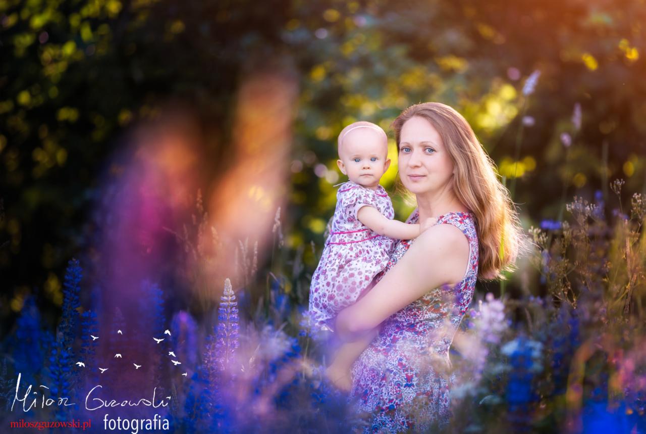 Kobieta trzymająca małą dziewczynkę w kwiecistej sukience na tle łąki z fioletowymi kwiatami, ciepłe światło słoneczne