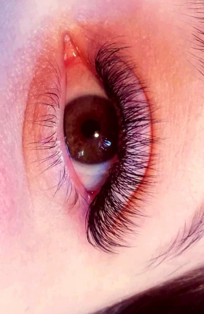 Zbliżenie na oko z brązową tęczówką i gęstymi, czarnymi, sztucznymi rzęsami, widoczne delikatne zaczerwienienie w kąciku oka.