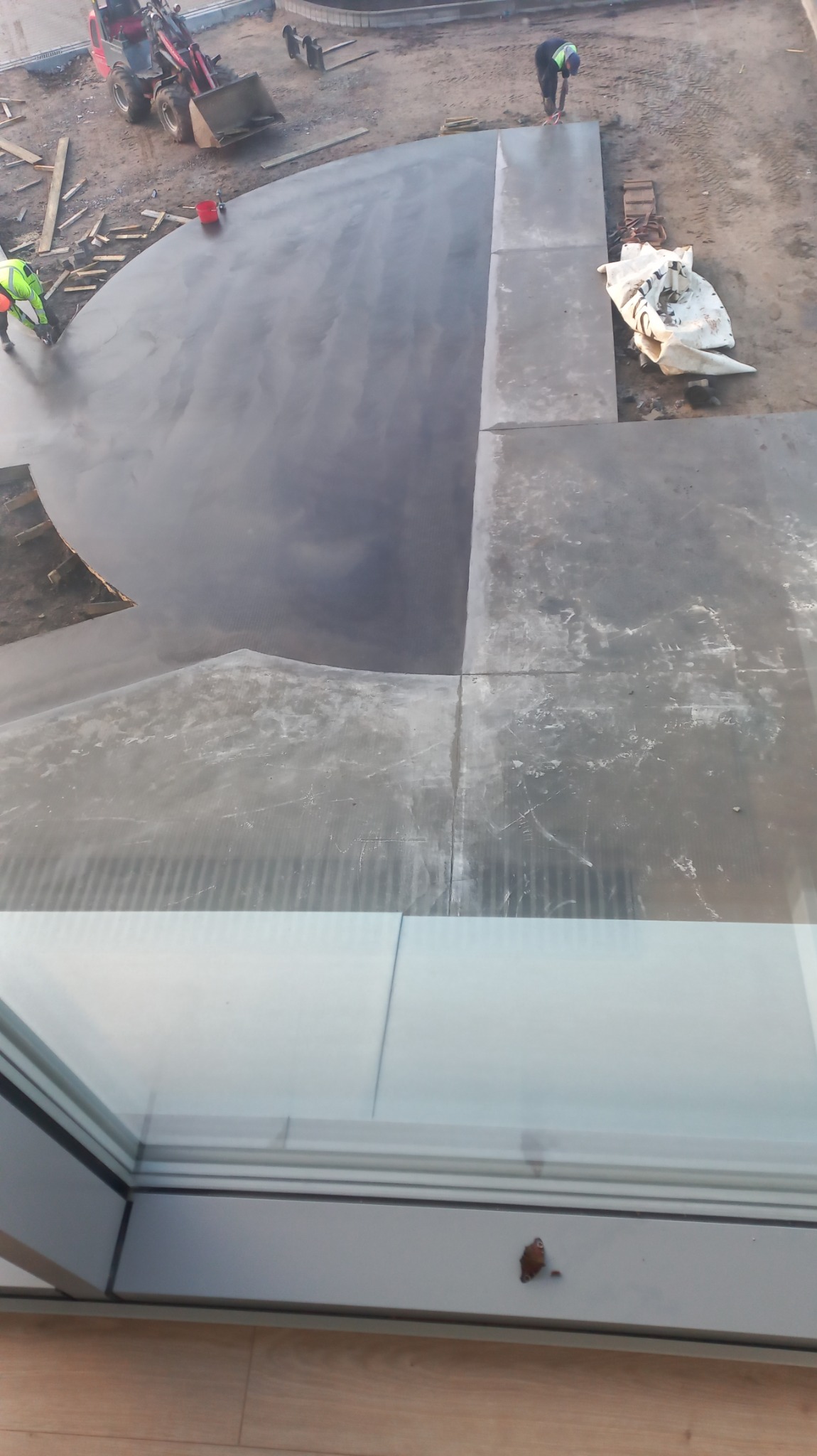 Widok z góry na wylewkę posadzki przemysłowej w kształcie półkola, z pracującym robotnikiem i minikoparką w tle, widziane przez okno z widocznym owadem na ramie.