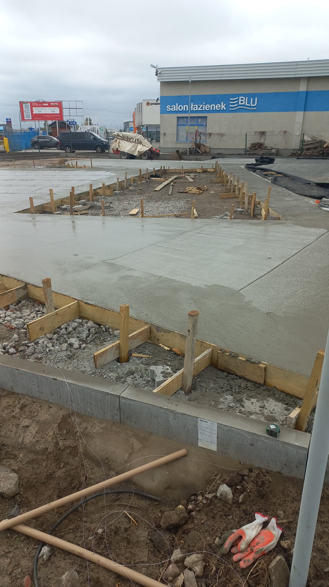 Wylewka betonowa z drewnianym szalunkiem i podsypką z kamieni, widoczne detale takie jak rękawice robocze i miarka zwijana przy krawężniku, w tle budynek salonu łazienek.