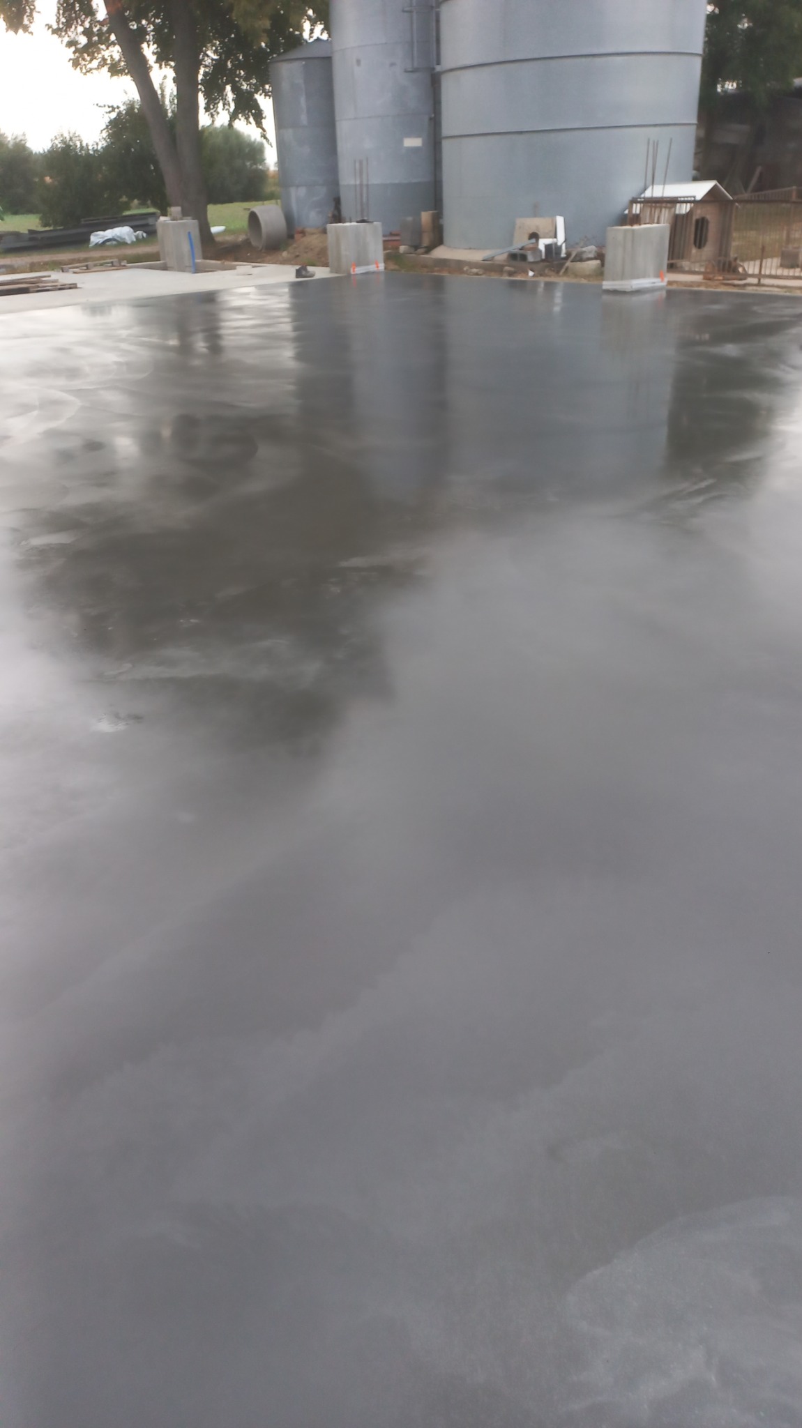 Świeżo wylana, mokra posadzka przemysłowa z widocznymi śladami po zacieraniu, na tle silosów zbożowych i elementów konstrukcyjnych.