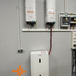 MULTISUN WIELKOPOLSKA SP.Z O.O. SP.K. - Biały magazyn energii z dwoma falownikami na ścianie, instalacja w szarym pomieszczeniu technicznym z widocznymi przewodami i zabezpieczeniami.