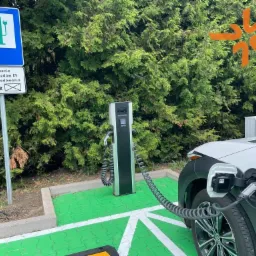 Biały samochód elektryczny ładowany przy stacji ładowania z zielonymi akcentami, na tle zielonych krzewów i znaku informacyjnego z symbolem ładowania EV.