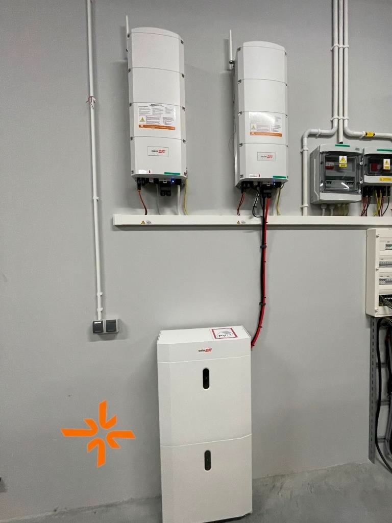 Biały magazyn energii z dwoma falownikami na ścianie, instalacja w szarym pomieszczeniu technicznym z widocznymi przewodami i zabezpieczeniami.