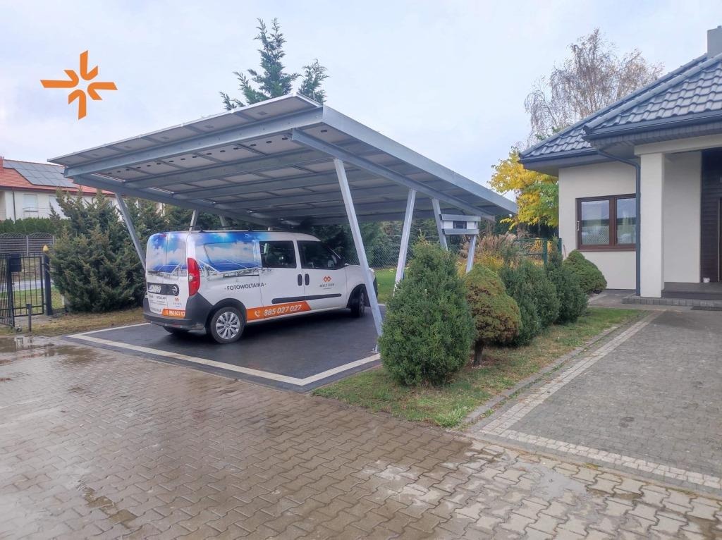 Zadaszenie carport z panelami słonecznymi nad białym samochodem dostawczym z brandingiem firmy, na tle domu jednorodzinnego.