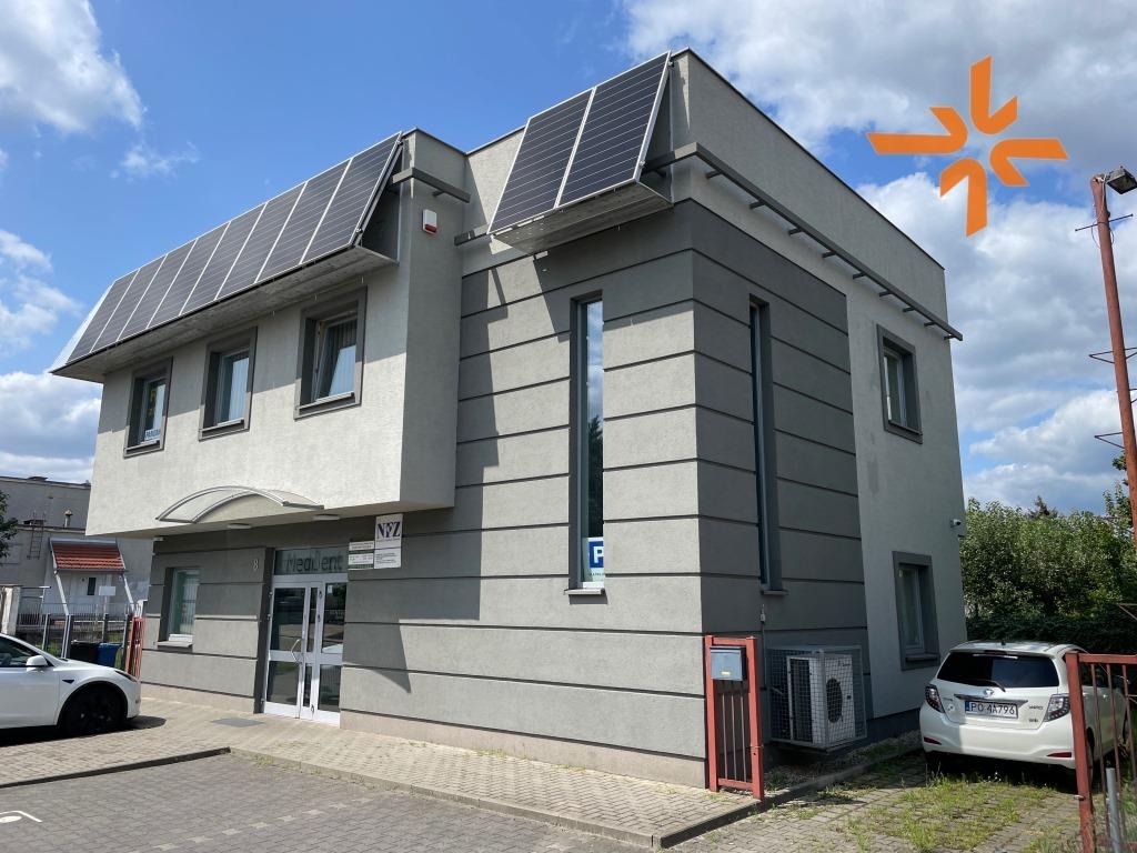 Dwukondygnacyjny budynek biurowy z panelami słonecznymi na dachu, widok z zewnątrz w słoneczny dzień. Biały samochód elektryczny i biały samochód spalinowy zaparkowane przed budynkiem.