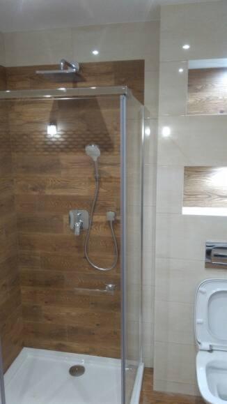 Kabina prysznicowa z drewnopodobnymi płytkami na ścianie, widoczny brodzik, bateria i deszczownica, obok toaleta w jasnej łazience.