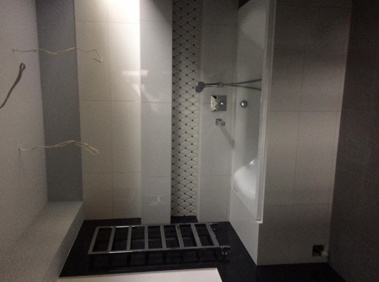 Nowoczesna łazienka w trakcie remontu: białe płytki, dekoracyjny pas mozaiki, czarne wykończenia podłogi, widoczne przewody elektryczne i metalowa konstrukcja na podłodze.