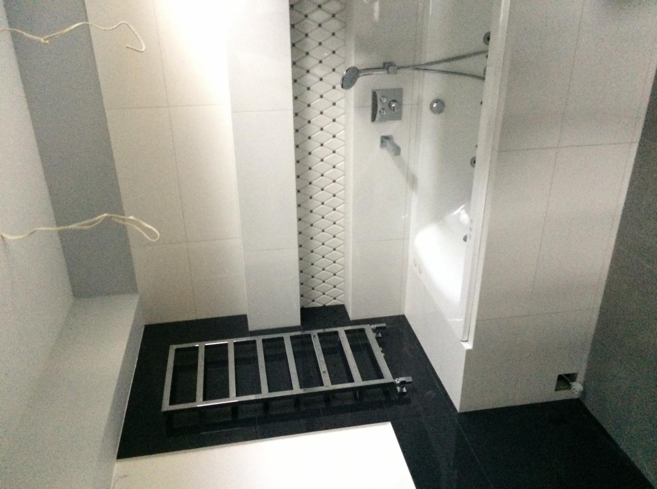 Wnętrze łazienki w trakcie remontu, widoczne płytki ścienne w odcieniach bieli i szarości, czarne płytki podłogowe oraz metalowa konstrukcja na podłodze i wystające przewody elektryczne ze ściany.