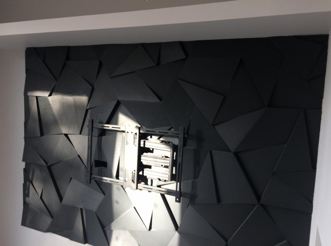 Czarna ściana z geometrycznym wzorem 3D, oświetlona z lewej strony, z zamontowanym uchwytem do telewizora.