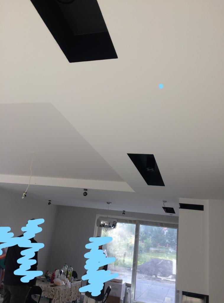 Biały sufit z czarnymi, prostokątnymi wnękami oświetleniowymi, widok na wnętrze pokoju z dużym oknem i częściowo zasłoniętymi osobami.