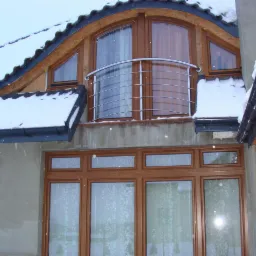 Fragment elewacji domu z oknami i balkonem zabezpieczonym metalową balustradą podczas opadów śniegu.