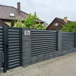 Nowoczesne ogrodzenie panelowe z poziomych lameli w kolorze grafitowym, z bramą wejściową, słupkami z szarej kostki, skrzynką gazową i numerem domu 12A przy ulicy Parkowej.
