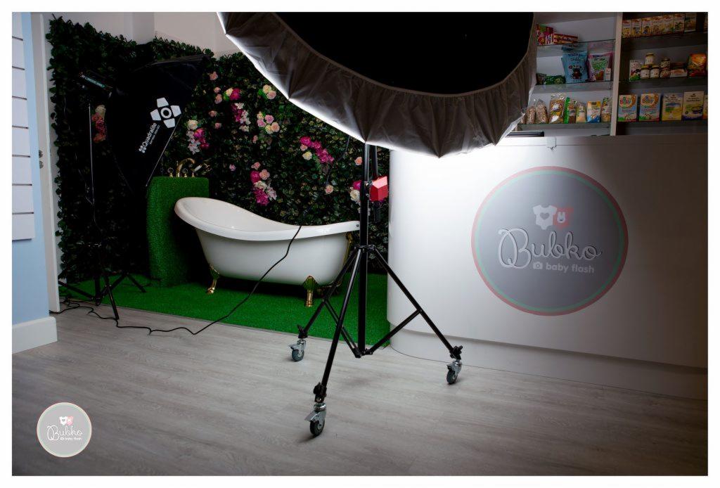 Przygotowane studio fotograficzne z wanną na złotych nóżkach, zielonym trawnikiem i ścianą z kwiatów, oświetlenie studyjne i logo 'Bubko baby flash'.