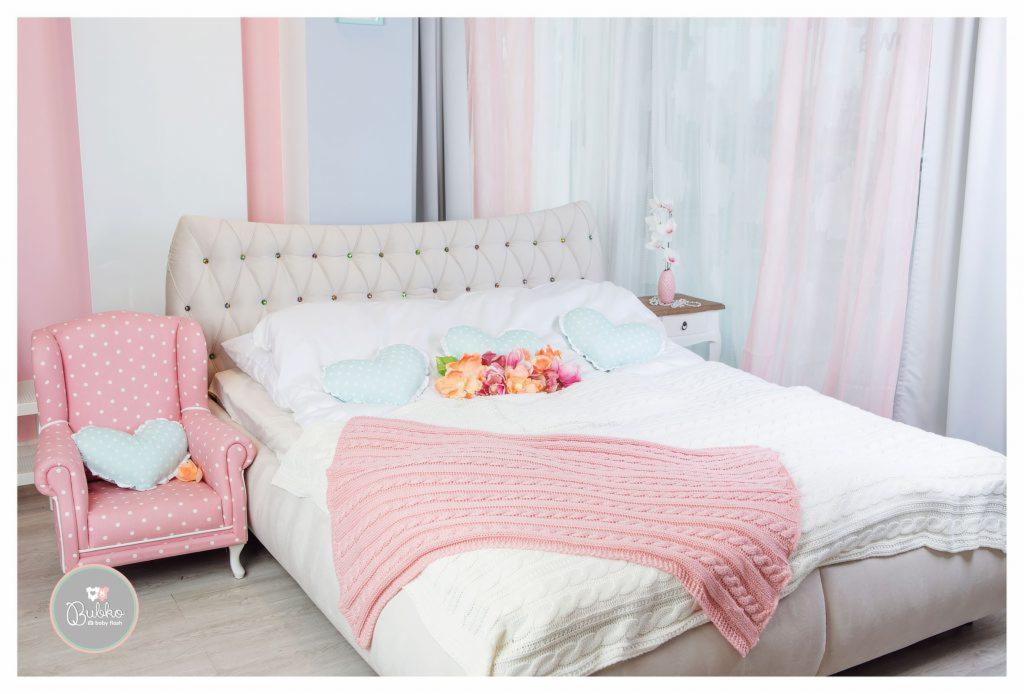 Jasny pokój z łóżkiem z pikowanym zagłówkiem, białymi poduszkami w kształcie serc, różowym pledem i bukietem kwiatów; obok różowy fotel w białe kropki z poduszką w kształcie serca.