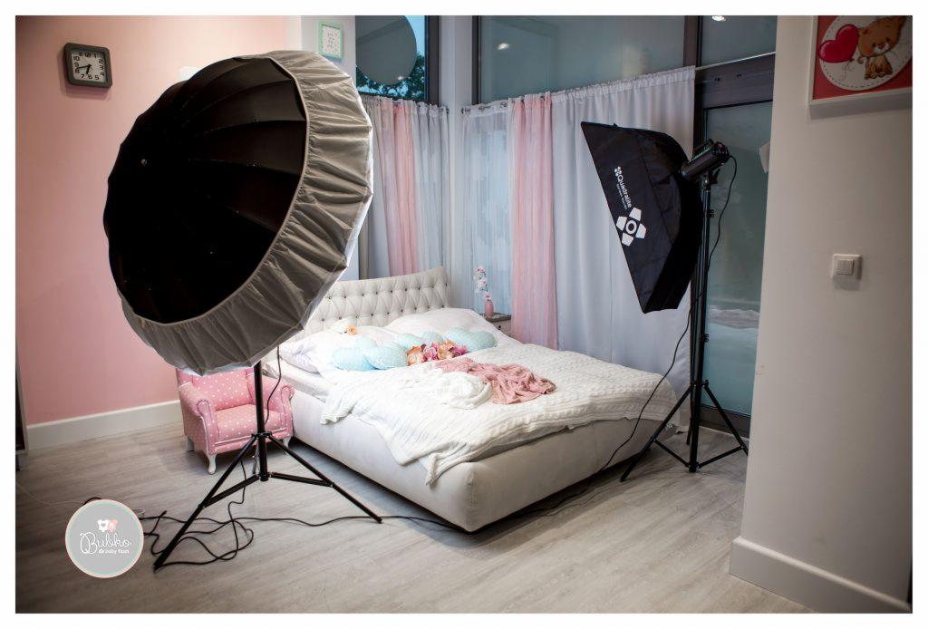 Przygotowane studio fotograficzne z lampami i softboxami, łóżko z dekoracjami w pastelowych kolorach, mały fotel dla dziecka i okno z delikatnymi zasłonami.