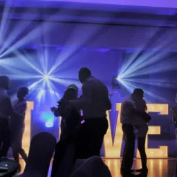 Tańczący goście weselni w niebieskim oświetleniu, duże podświetlane litery tworzące napis za nimi, reflektory na scenie.