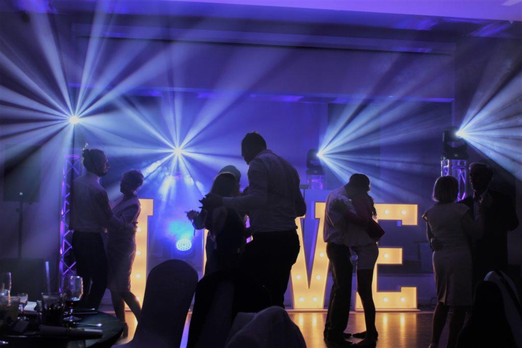 Tańczący goście weselni w niebieskim oświetleniu, duże podświetlane litery tworzące napis za nimi, reflektory na scenie.