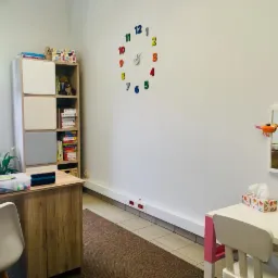 Gabinet terapeutyczny z biurkiem, regałem z książkami i zabawkami edukacyjnymi, kolorowym zegarem na ścianie i stolikiem dla dzieci z chusteczkami.