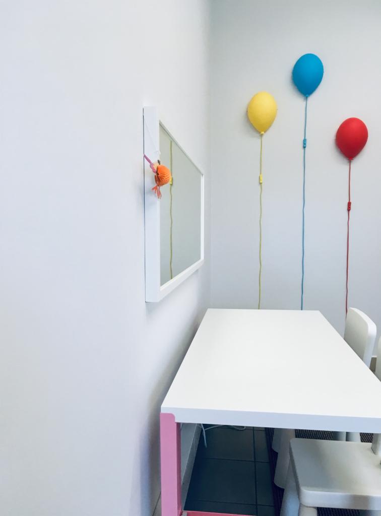 Jasny pokój z białym stołem, krzesłami, lustrem z pomarańczową ozdobą i trzema lampami ściennymi w kształcie balonów: żółtym, niebieskim i czerwonym.