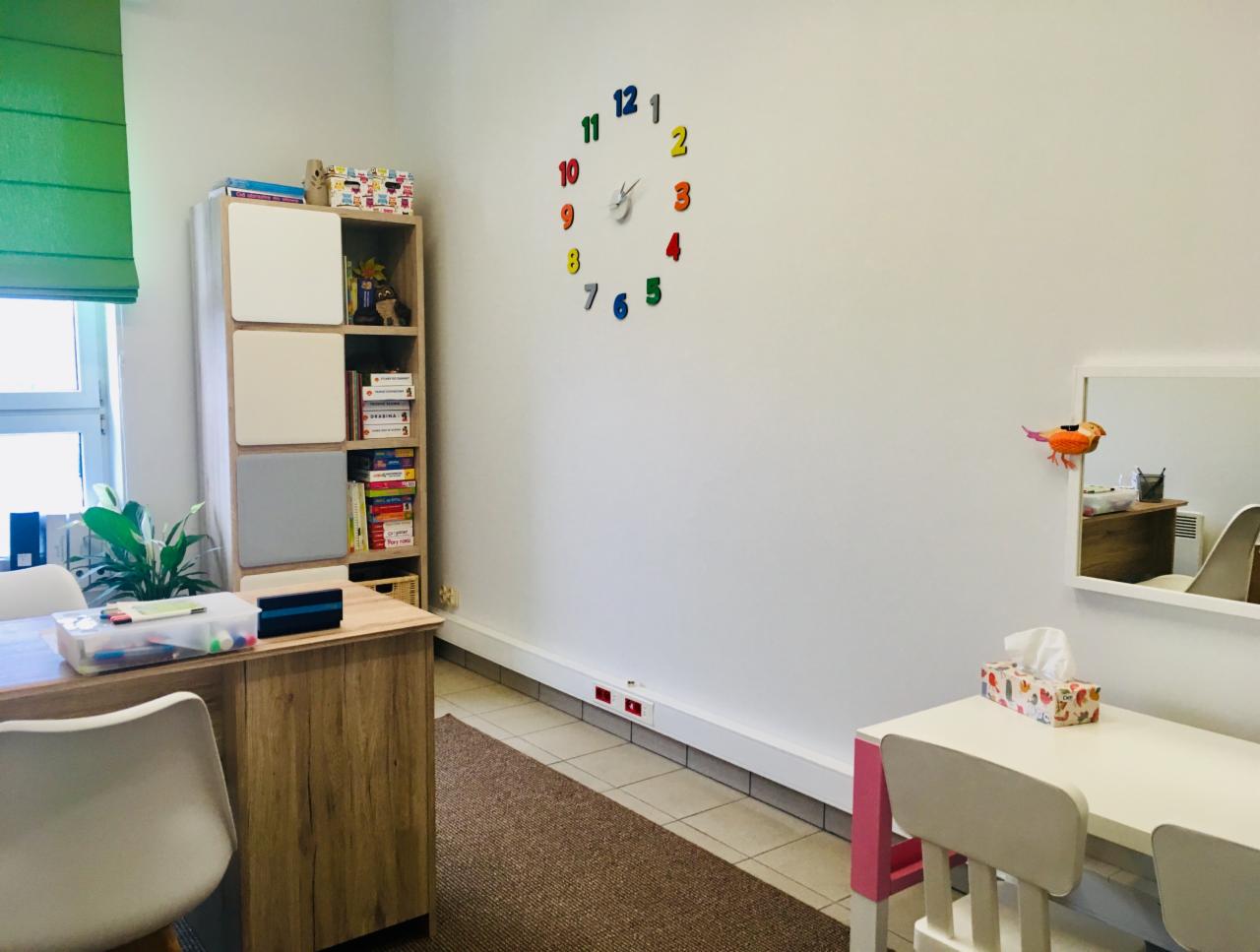 Gabinet terapeutyczny z biurkiem, regałem z książkami i zabawkami edukacyjnymi, kolorowym zegarem na ścianie i stolikiem dla dzieci z chusteczkami.