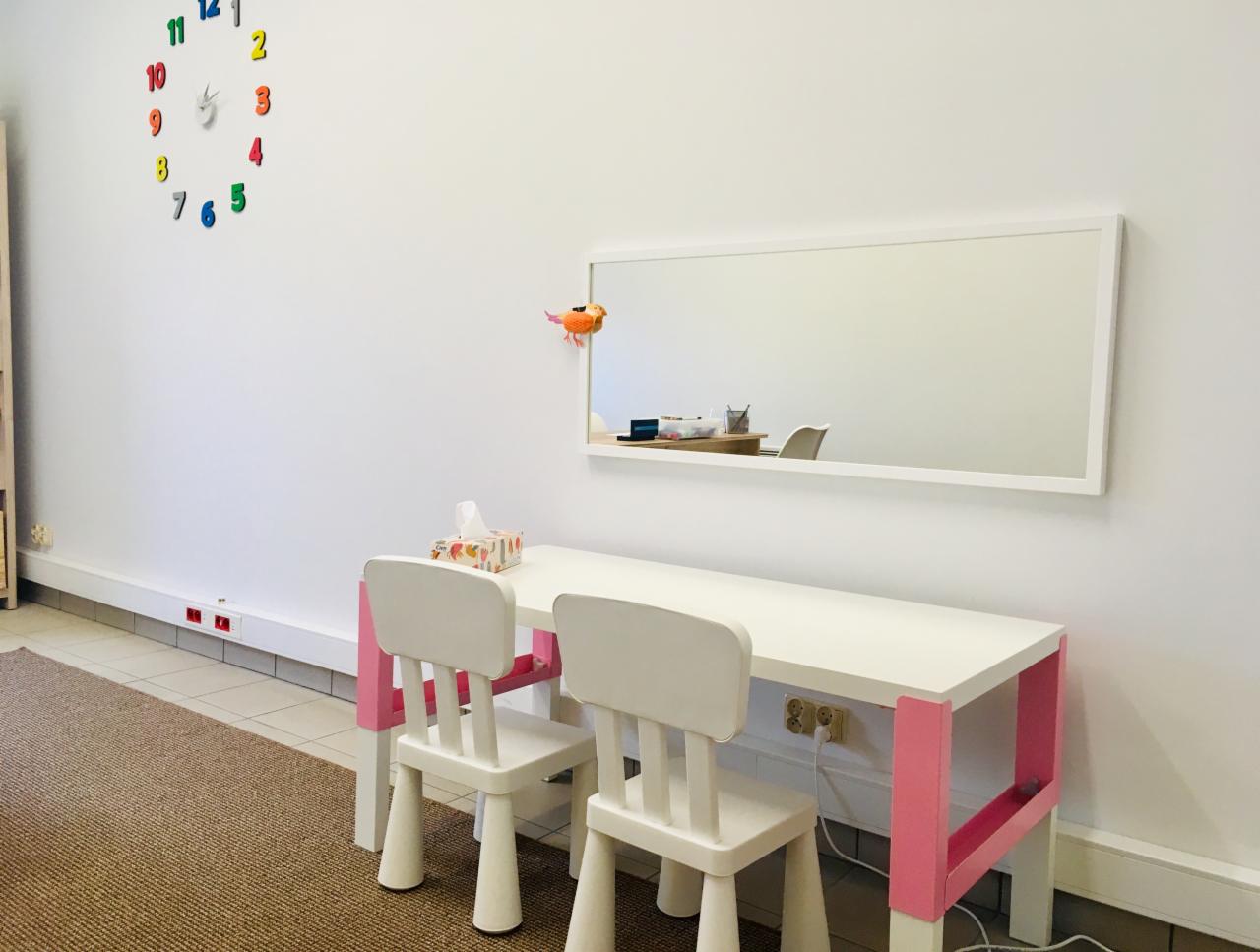 Jasny pokój z białym stolikiem z różowymi nogami i dwoma krzesłami, lustro na ścianie, kolorowy zegar z cyframi i dywan na podłodze.