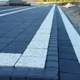 Nowo ułożony chodnik z ciemnoszarej kostki brukowej z białymi pasami po bokach i wzdłuż, widoczne krawężniki i fragment terenu przygotowanego pod dalsze prace.