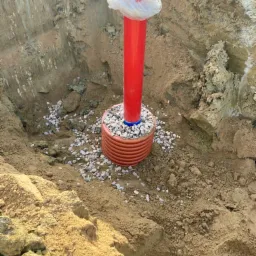 Czerwona rura wystająca z wykopu, zabezpieczona folią, otoczona drenażem z małych kamieni.