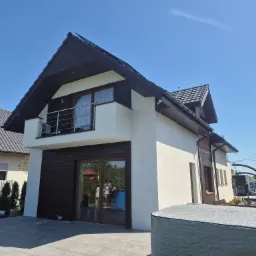 Nowoczesny dom jednorodzinny z jasną elewacją i ciemnobrązową drewnianą okładziną, widoczne okna i balkon z metalową barierką, na pierwszym planie przykryty basen ogrodowy.