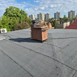 Pokrycie dachu papą termozgrzewalną na płasko, z widocznym kominem wentylacyjnym i w tle budynki mieszkalne.