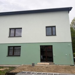 MARKO FIRMA BUDOWLANA - Dwukolorowa elewacja domu po remoncie: dolna część zielona, górna biała. Widoczne okna z brązowymi ramami i drzwi wejściowe. Na pierwszym planie ziemia i drabina.