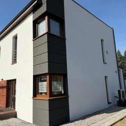 MARKO FIRMA BUDOWLANA - Nowoczesny dom z białą elewacją i grafitowymi panelami narożnymi, drewnianymi oknami i tarasem. Widok z zewnątrz, słoneczny dzień.