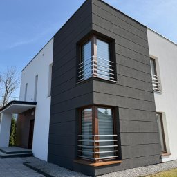 MARKO FIRMA BUDOWLANA - Dwukolorowa elewacja domu jednorodzinnego z grafitową częścią z poziomymi pasami i białym tynkiem. Okna z balustradami. Widok z zewnątrz, w słoneczny dzień.