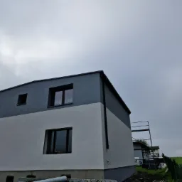 Dwukolorowa elewacja domu jednorodzinnego z widocznymi rusztowaniami i pochmurnym niebem w tle.