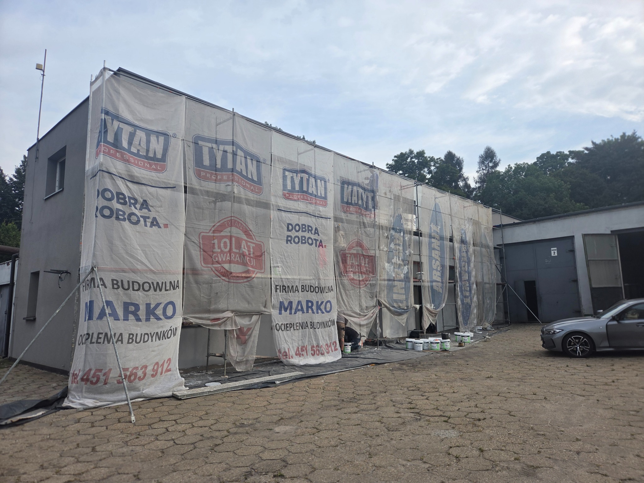 Budynek w trakcie prac, osłonięty siatką z reklamami. Widoczne farby, narzędzia i pracownik. Szara elewacja. Częściowo zasłonięty budynek w trakcie ocieplania.