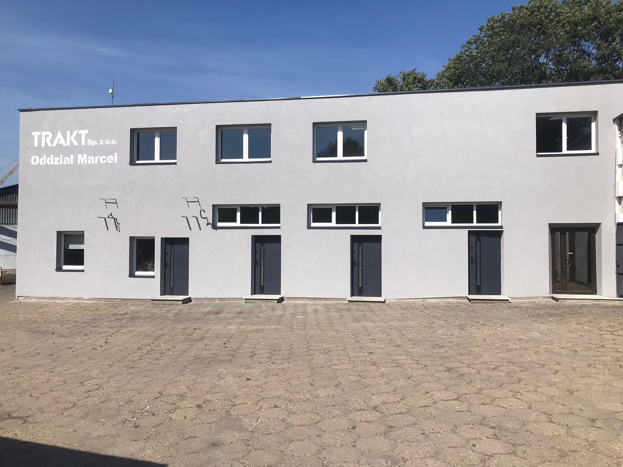 Szary budynek biurowy z białymi oknami i ciemnymi drzwiami wejściowymi. Widoczne logo firmy 'TRAKT' na elewacji. Minimalistyczny styl, prosta bryła budynku.