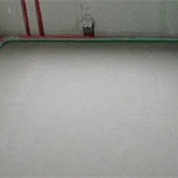 Świeżo wylana, gładka posadzka jastrychowa w pomieszczeniu, z widocznymi rurami grzewczymi w izolacji przy ścianach i elementami instalacji wodnej na ścianie.