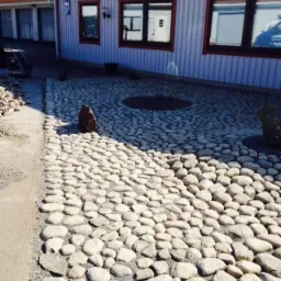 Ułożona ścieżka z jasnych, okrągłych kamieni prowadząca do budynku z szarą elewacją i czerwonym dachem; w tle widoczne garaże.