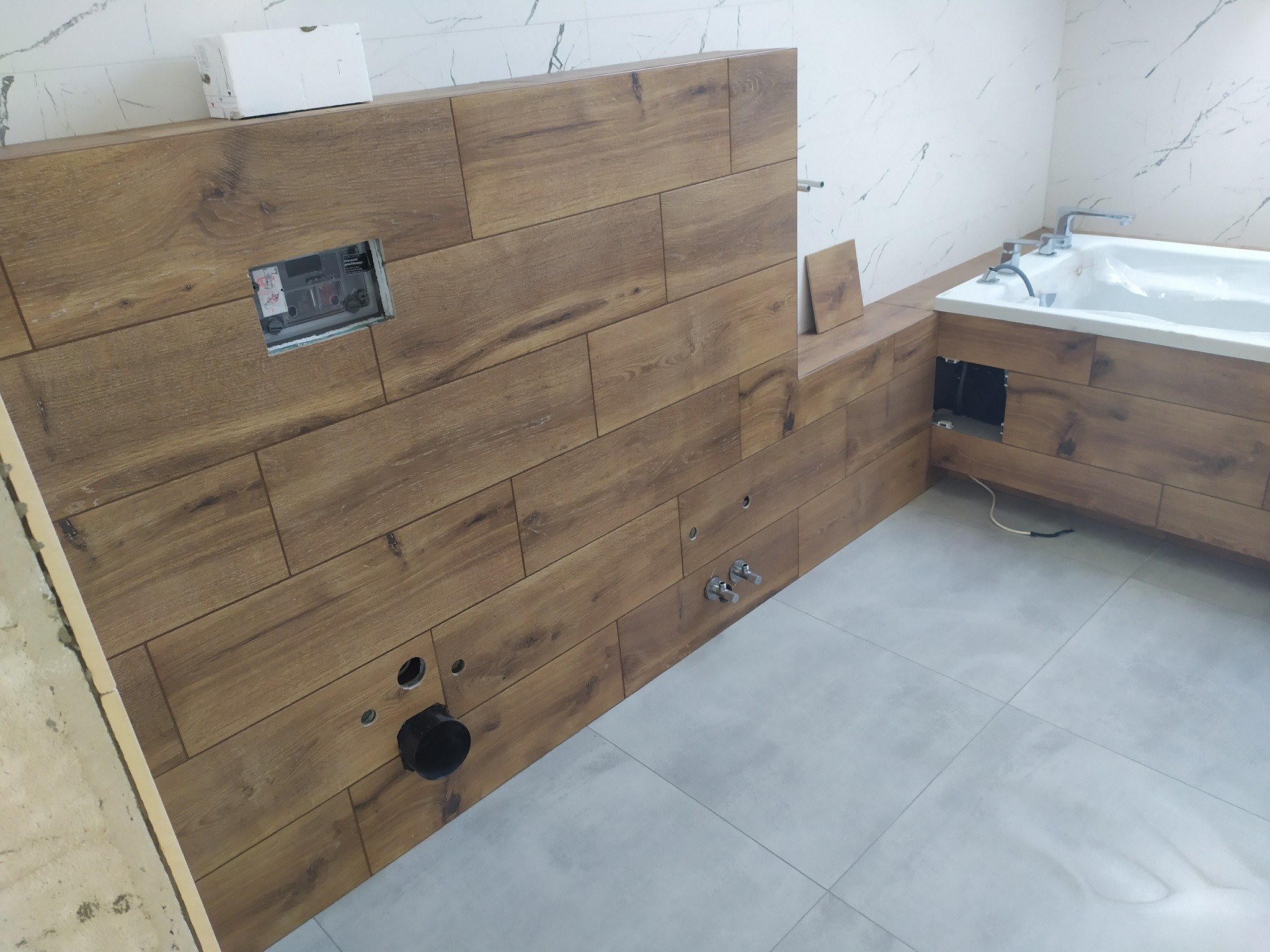 Ściana obudowy wanny wyłożona płytkami imitującymi drewno, z widocznymi otworami na instalacje i niezamaskowaną puszką elektryczną, szare płytki podłogowe, białe marmurowe płytki na ścianach w tle.