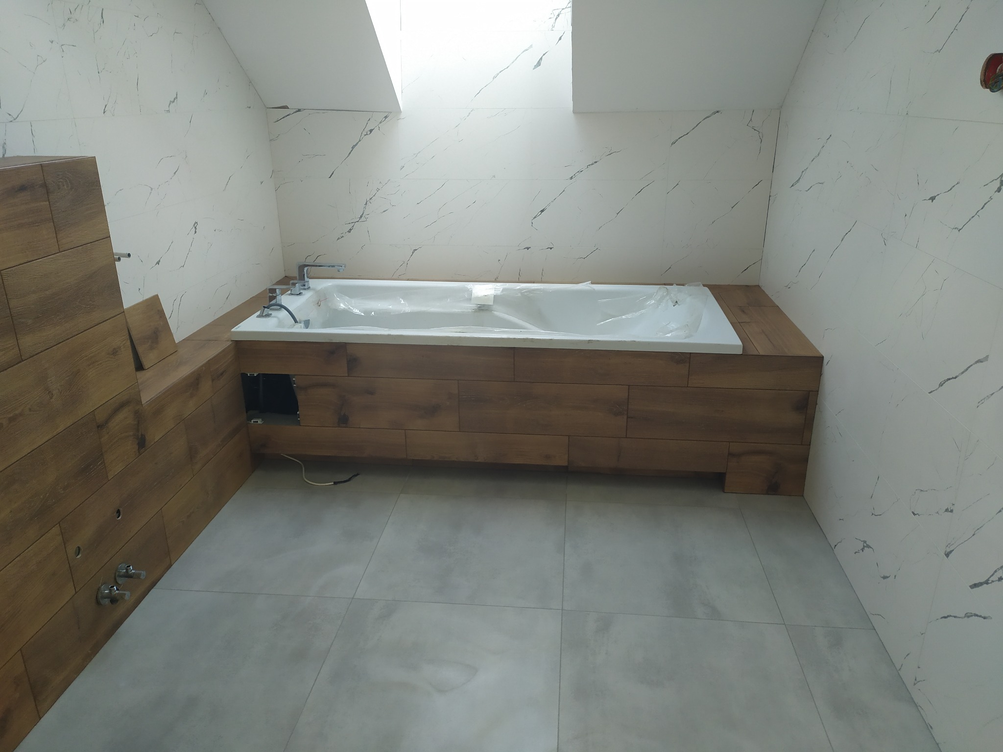 Nowoczesna łazienka w trakcie wykańczania: białe płytki marmurowe na ścianach, wanna obudowana drewnem, szare płytki podłogowe, okno dachowe w skosie.