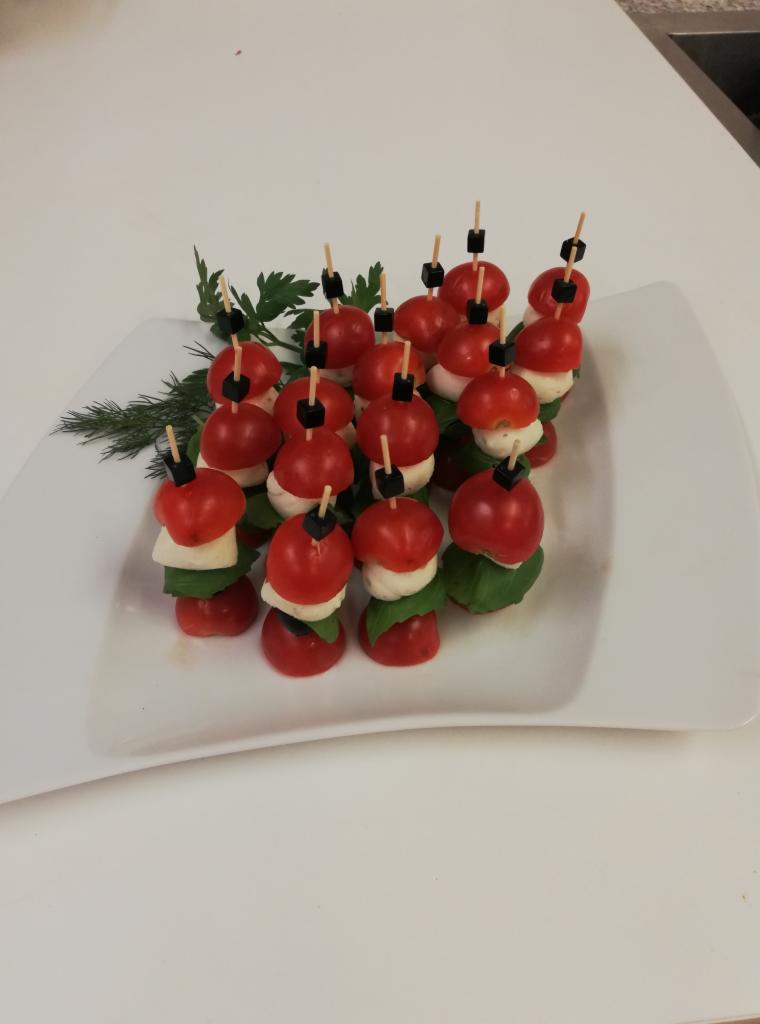Elegancko ułożone koreczki Caprese na białym talerzu, ozdobione zielonymi gałązkami, z widocznymi warstwami pomidorków cherry, mozzarelli i bazylii, spięte wykałaczkami z czarnymi oliwkami.