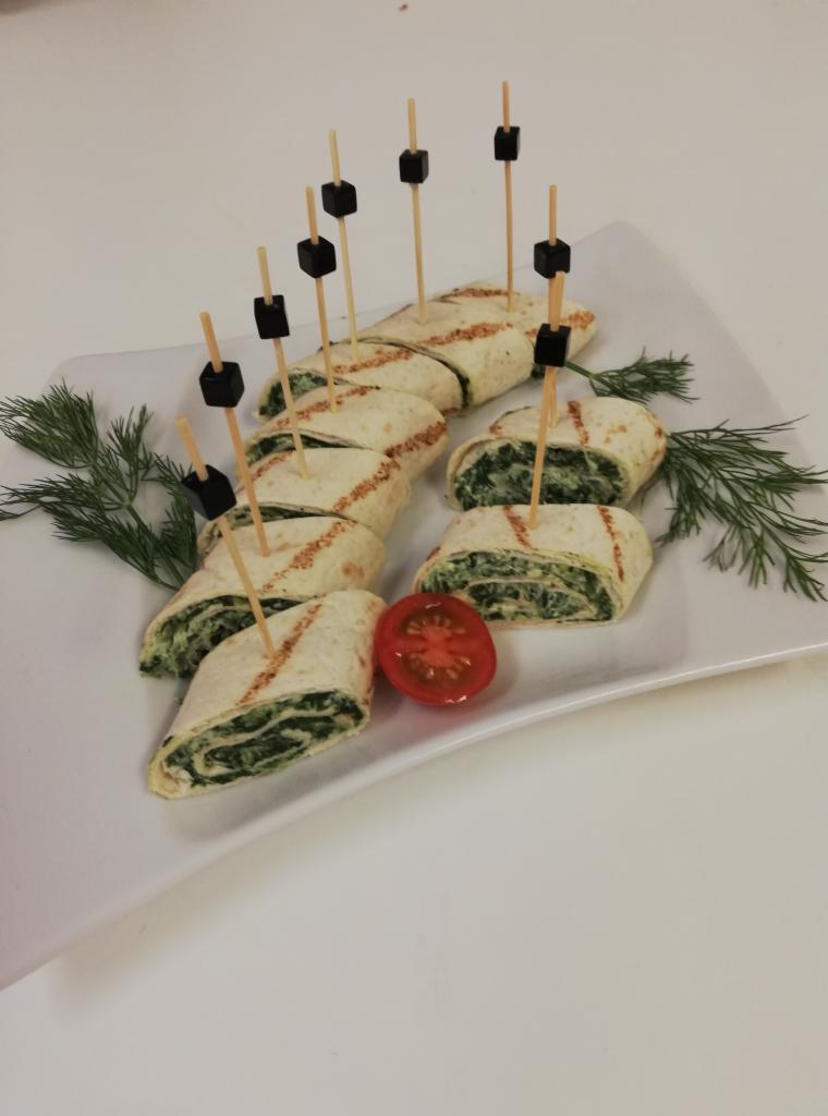 Tortilla wrap z nadzieniem szpinakowym, przekrojona na kawałki i spięta wykałaczkami z czarnymi kostkami, ułożona na białym talerzu z gałązkami koperku i połówką pomidora.