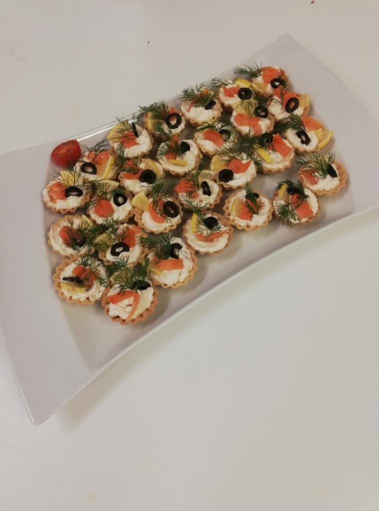 Talerz z kruchymi babeczkami wypełnionymi kremowym serem, łososiem, plasterkami cytryny, oliwkami i koperkiem, uzupełniony pojedynczym pomidorkiem koktajlowym.