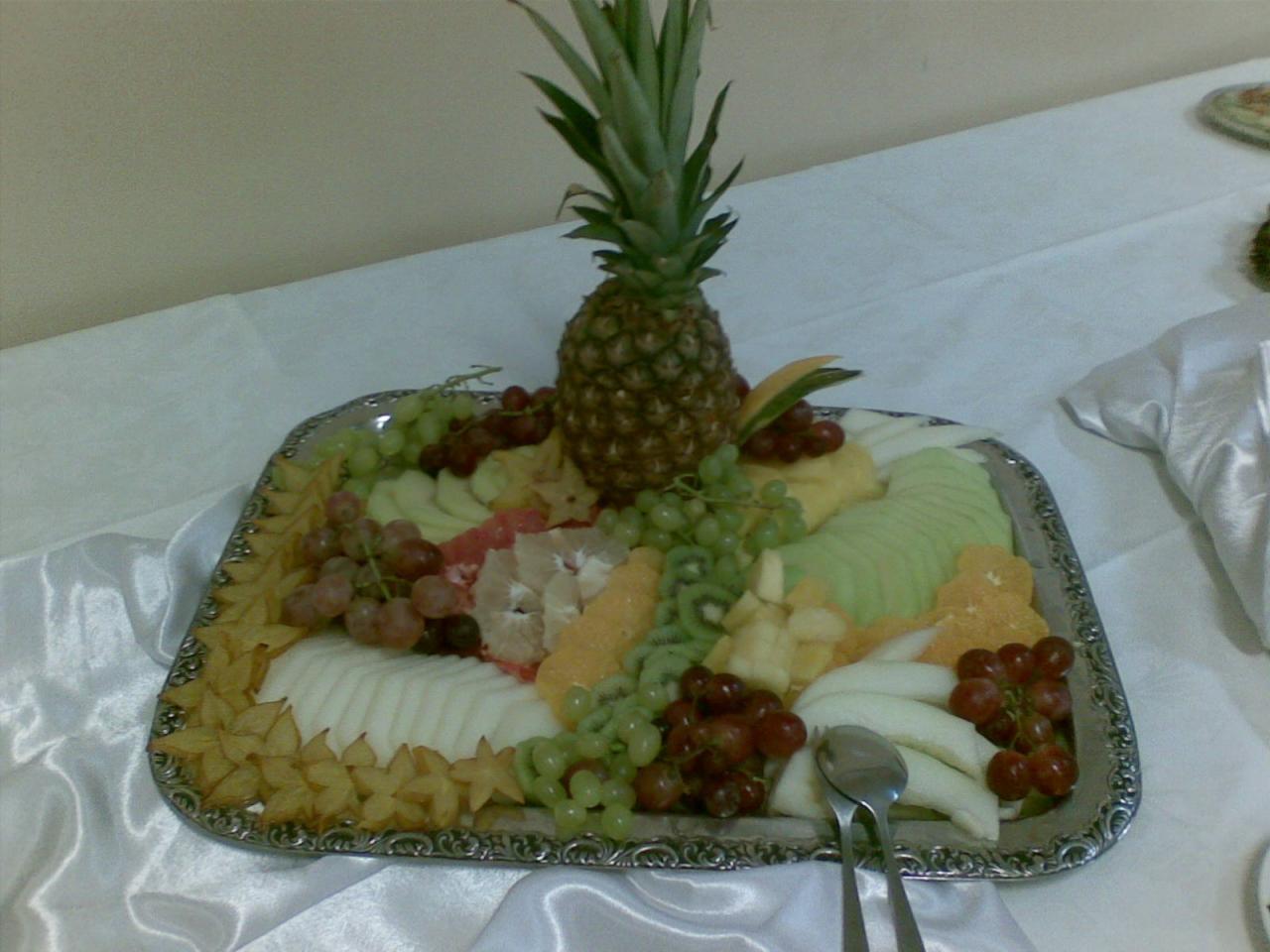Srebrna patera z kompozycją z pokrojonych owoców: ananas, winogrona, melon, kiwi, karambola, pomarańcze, banany, na białym obrusie, obok leżą dwie łyżeczki.
